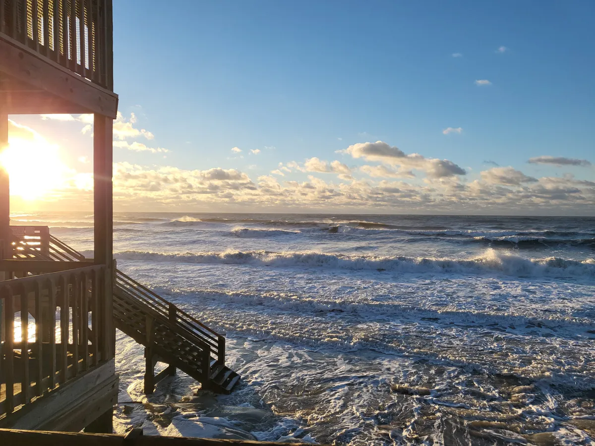 A stunning sunset over the ocean, waves lap against the shore, and a wooden structure with stairs offers direct access to the sand. The scene is calm, inviting residents to enjoy tranquil moments and the salty breezes described in the section.