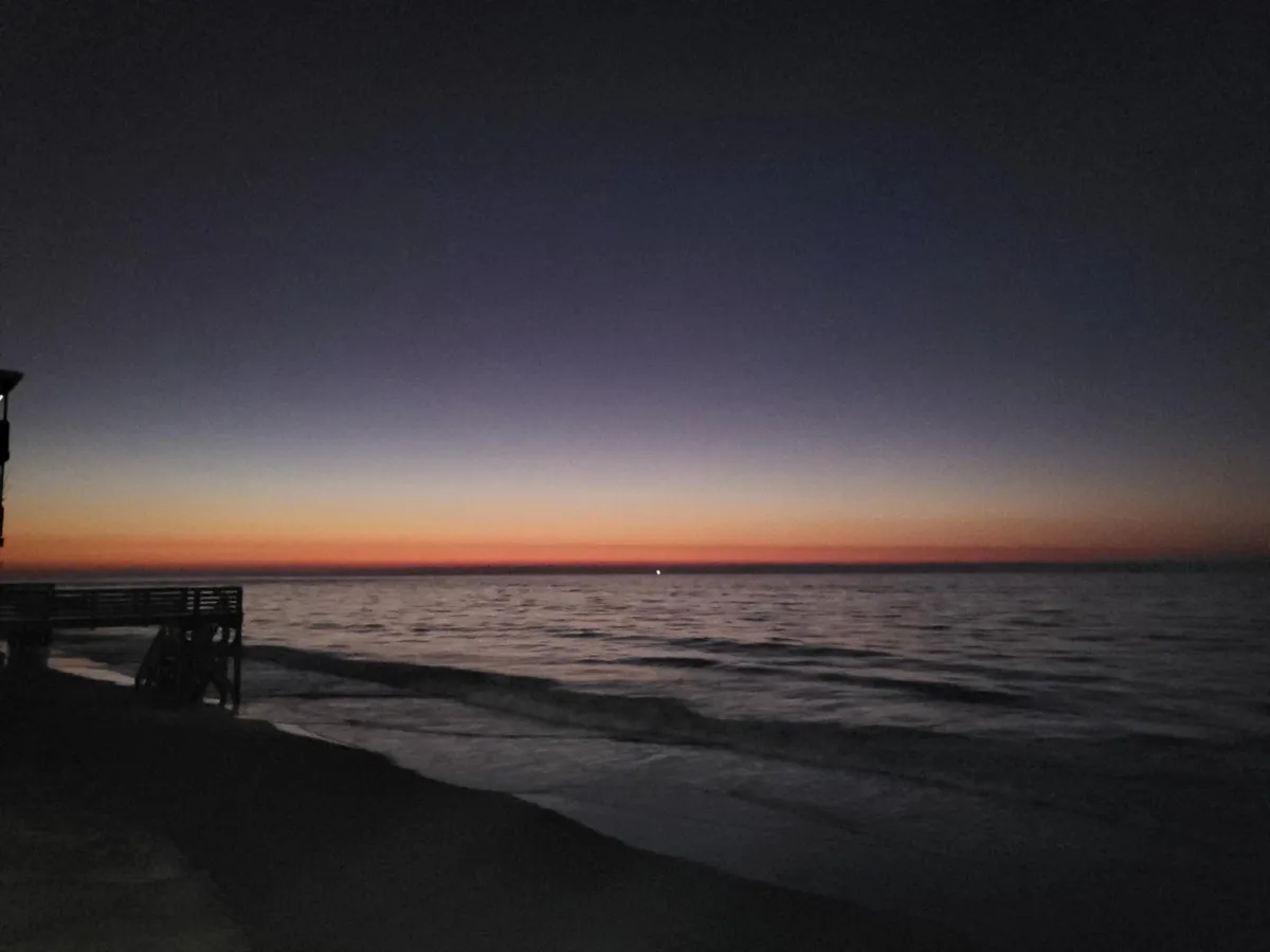 A serene sunset unfolds over the ocean, with gentle waves reflecting the vibrant colors of dusk. A pier extends into the water, inviting evening strolls, quiet moments of reflection, and opportunities for photography. The tranquil scene captures the changing moods of the coast, from energetic days to peaceful nights.