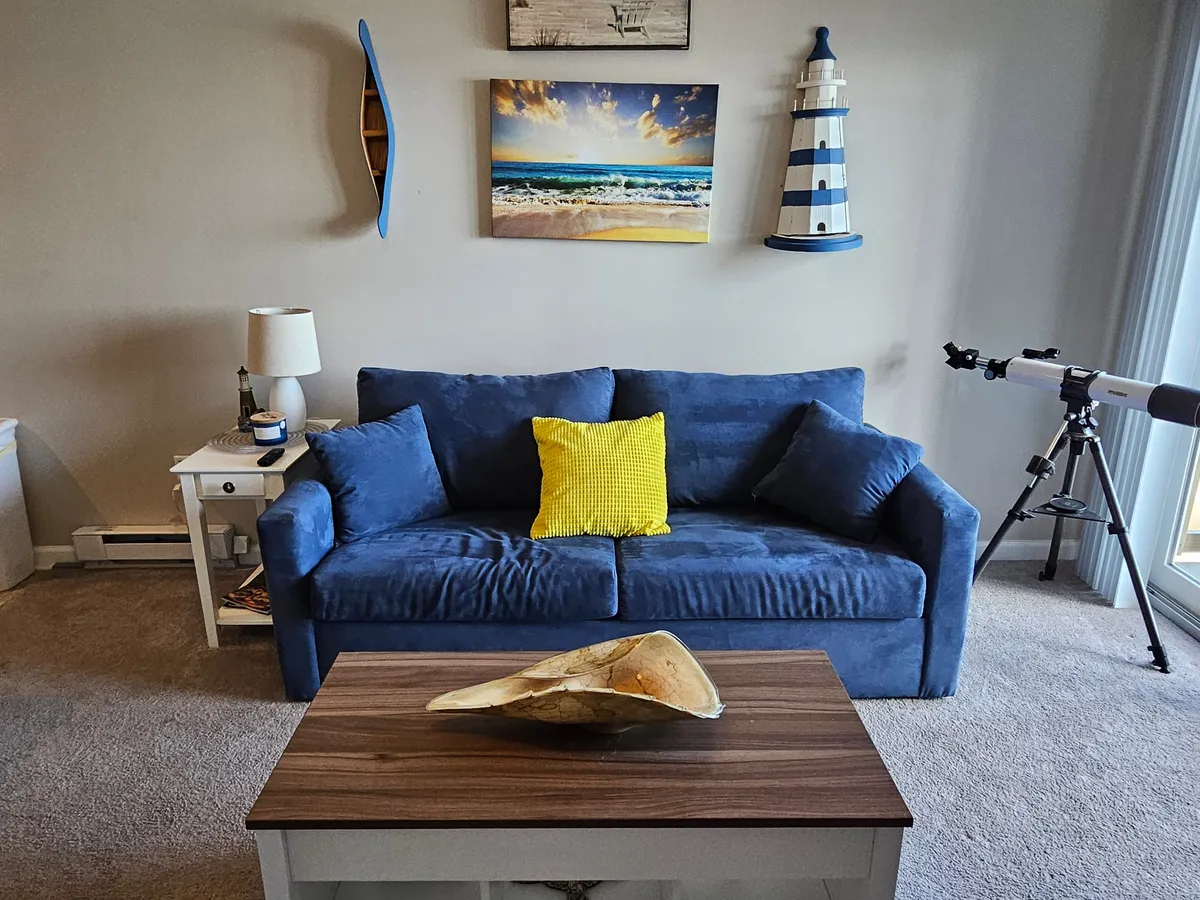 A tranquil and cheerful living room featuring a blue sofa adorned with a yellow pillow, beach scene artwork, lighthouse decor, and a telescope. The space is filled with natural light and includes a coffee table for gatherings.