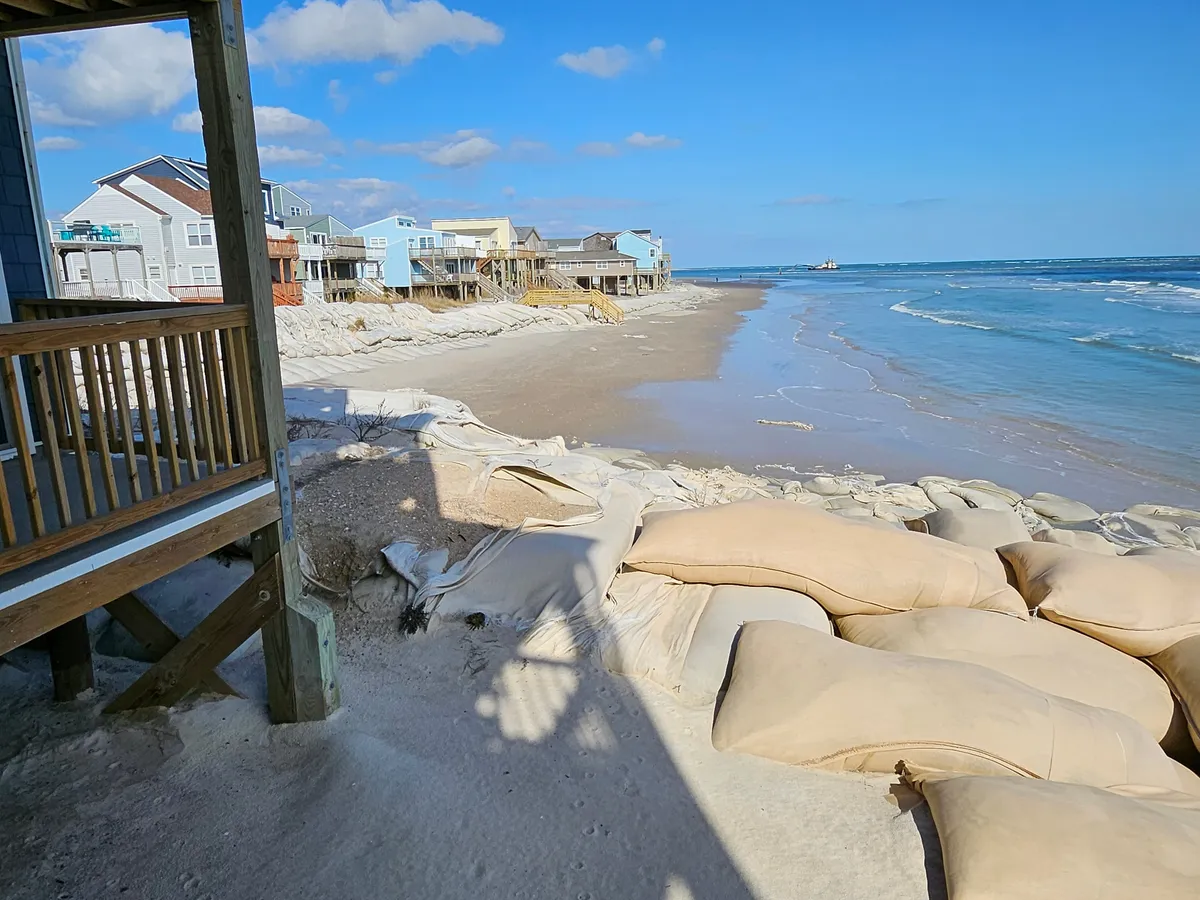 A serene beachside view with rows of beachfront houses, gentle ocean waves, scattered sandbags, and a pier visible in the distance. The setting is perfect for beachcombing, fishing, and scenic walks.