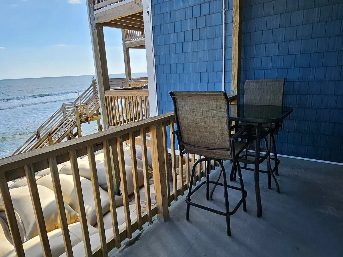 A private balcony with a wooden railing overlooks the sparkling Atlantic, set up with a table and chairs perfect for enjoying breakfast or coffee while listening to the waves. The scene exudes calm and tranquility, with the ocean just steps away.