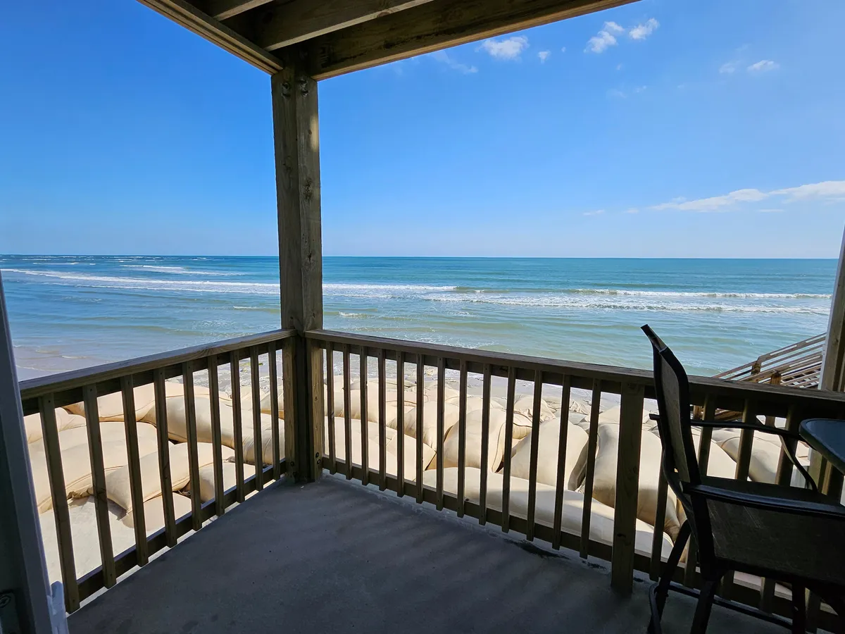 A serene oceanfront balcony scene with a wooden railing overlooking the sandy beach and the Atlantic Ocean. The sand and water are just steps away, emphasizing the direct connection to the shore. Gentle sunlight and a tranquil atmosphere invite relaxation and enjoyment of the views.