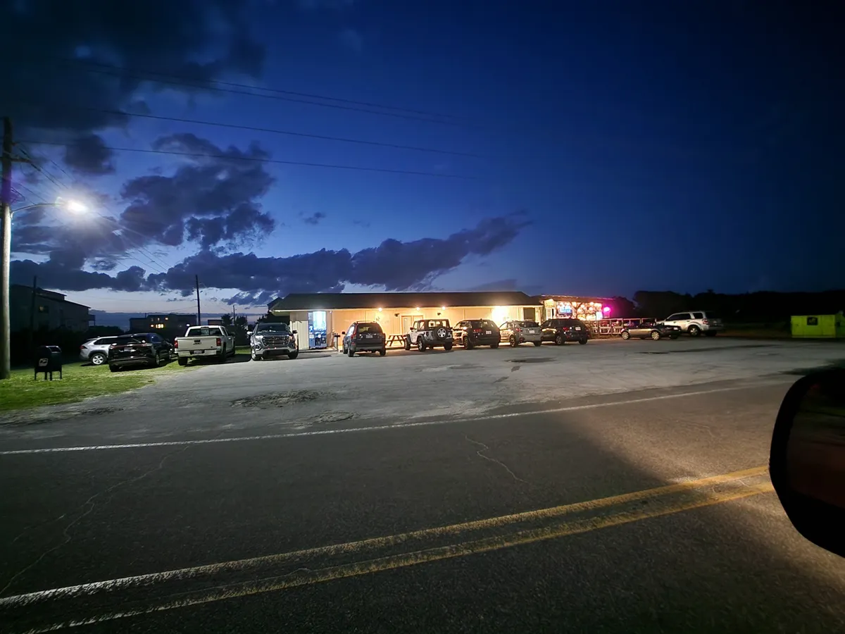 The building is warmly lit at night, with parked cars and streetlights creating a sense of welcoming activity. The scene hints at social gatherings, friendly interactions, and the vibrancy that continues after sunset. The illuminated exterior symbolizes comfort and community in the evenings, regardless of the time of year.