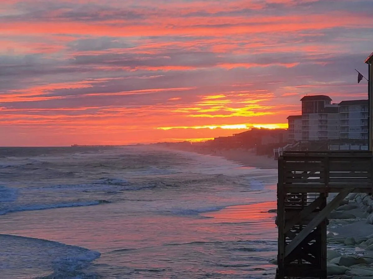 A breathtaking view of the sun setting over the ocean, with a pier stretching into the water and building silhouettes on the coast. The tranquil scene suggests peaceful moments and vibrant social activity during the golden hour.
