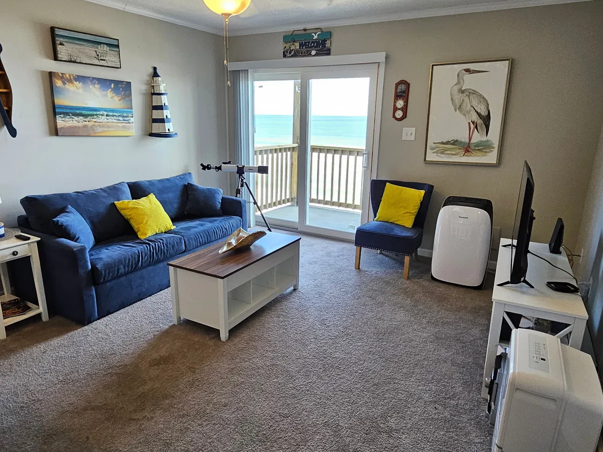 A cozy blue sofa sits beside a coffee table and a telescope, facing large sliding glass doors that open to the balcony and the ocean beyond. The inviting setup is ideal for enjoying the view, reading, or stargazing from the comfort of the living area.