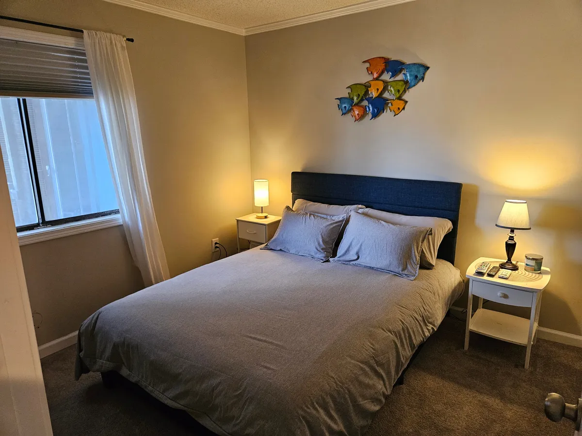 A serene bedroom scene featuring a comfortable bed with light bedding, a distinctive fish-themed wall art, a nightstand with a lamp, and soft natural lighting. The space is designed for relaxation and evokes a tranquil, coastal vibe.