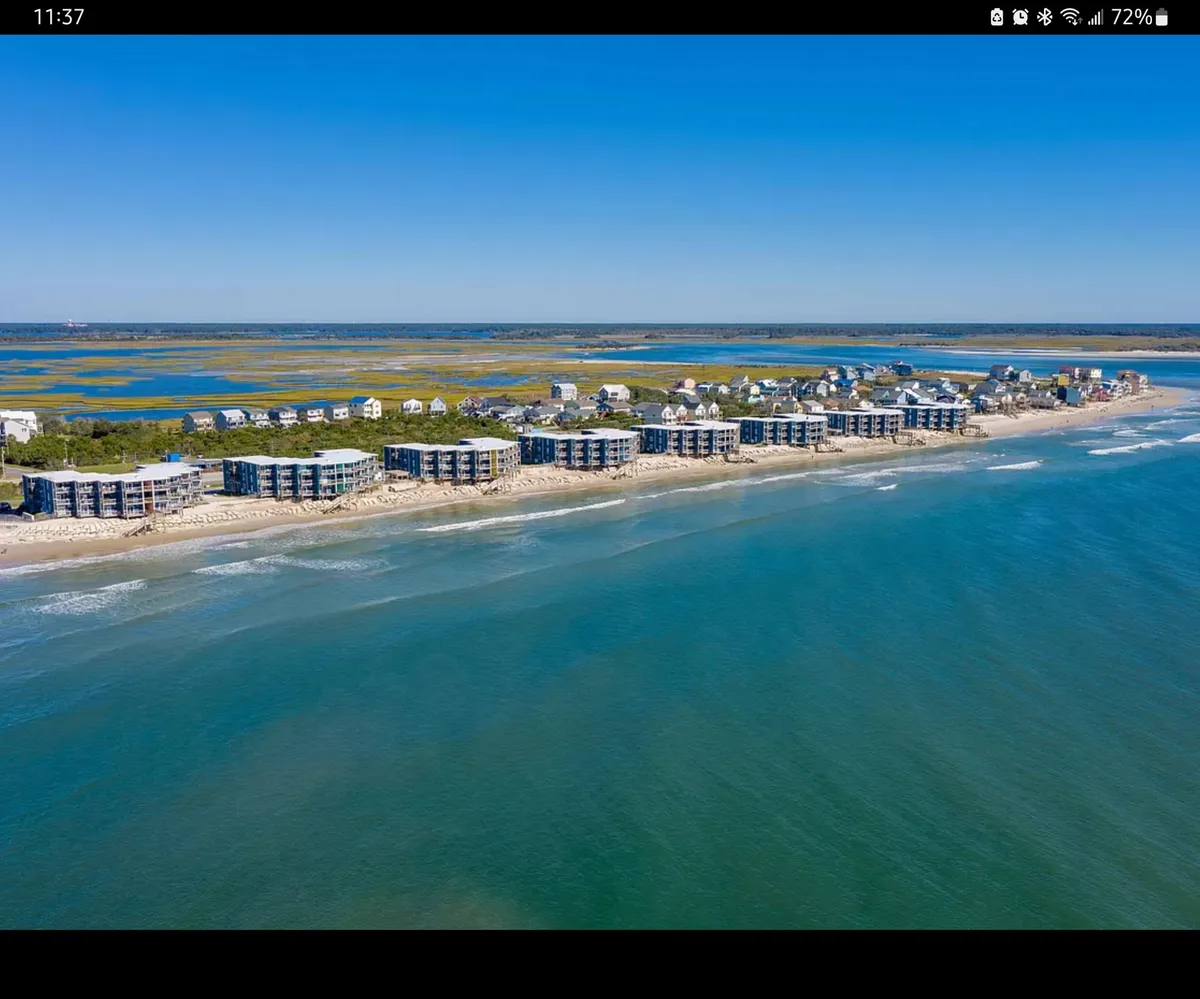 A wide exterior shot of beachfront property, ocean waves, lush green marshlands, and residential buildings. The image captures the broader setting, showing how close the building sits to the water and framing the uninterrupted access to the shoreline.