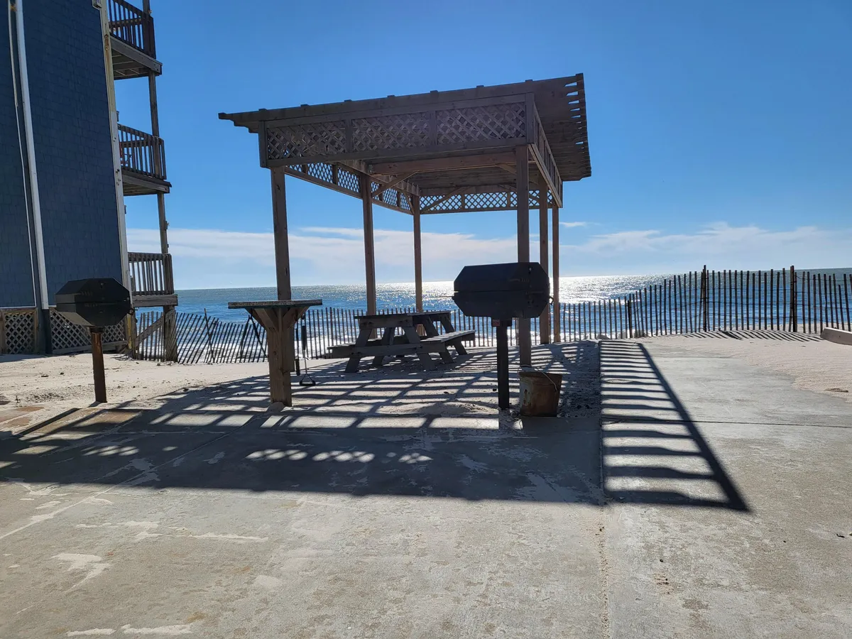 A welcoming picnic area sits beside the ocean, with a sturdy wooden table and grill arranged for outdoor meals. Gentle sunlight casts patterned shadows across the space, while the expansive sea view suggests a relaxing and social atmosphere. This communal setting is ideal for enjoying meals, sunset views, and time with friends or family, evoking the pleasures of outdoor living in North Topsail Beach across the seasons.
