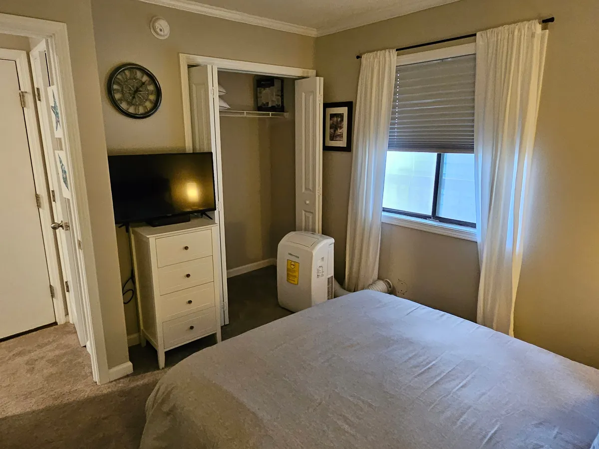 A cozy bedroom setup featuring a bed, a television positioned for easy viewing from bed, a window letting in natural light, a matching dresser, and an air conditioner. The space feels inviting and modern, tailored for relaxation or entertainment.