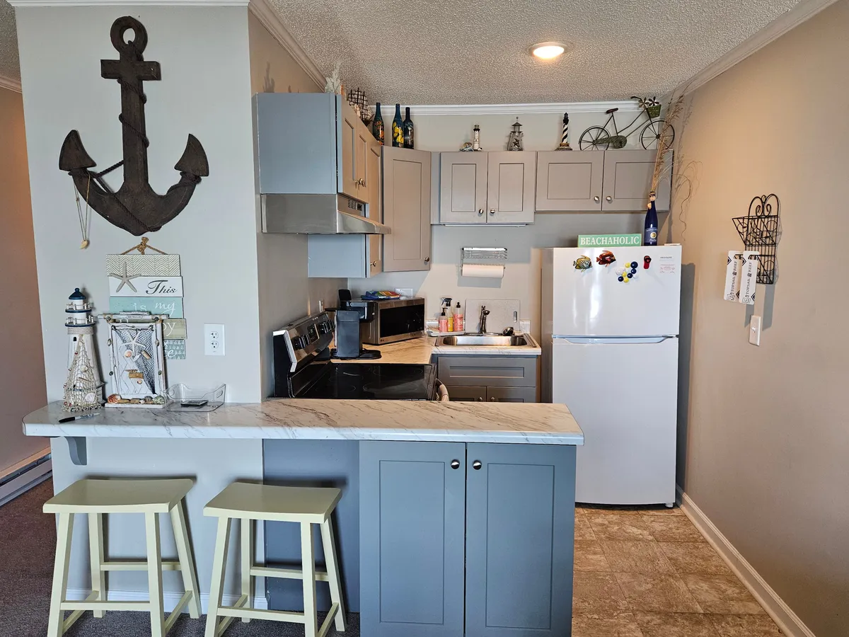 A cheerful, light-filled kitchen scene featuring anchor decor above the cabinetry, bar stools at the counter, modern kitchen appliances, and decorative bottles on display. The space is designed for both cooking and socializing, with a welcoming atmosphere.