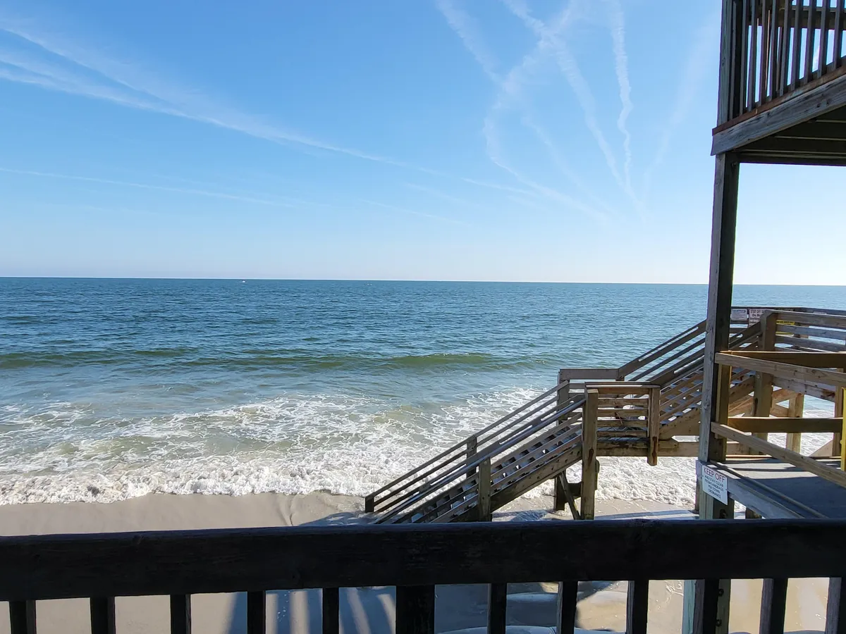 The hero image showcases a stunning first-floor oceanfront view, with gentle stairs leading directly from a private balcony onto the sandy beach. The rolling waves, golden sunset hues, and the inviting shoreline create a tranquil and welcoming scene, perfect for beach activities and relaxation.