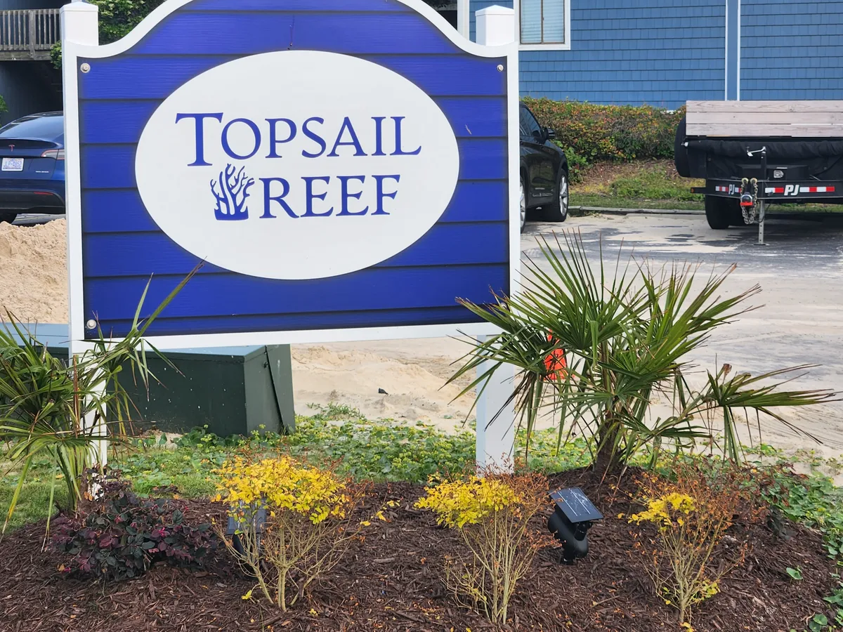 The entrance to the Topsail Reef community is framed by lush, well-maintained landscaping and a welcoming sign. The environment conveys a sense of arrival and hints at a friendly, resort-inspired setting with easy access to local attractions.