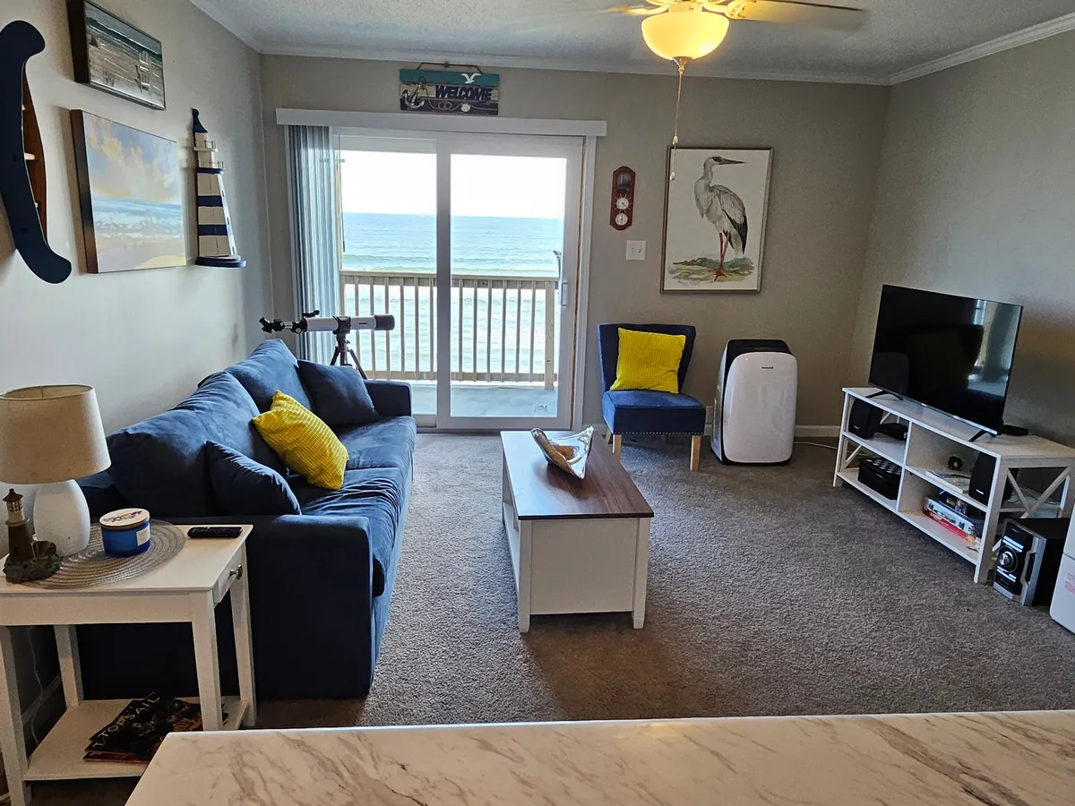 A bright, cozy living area featuring a blue sofa, coffee table, wall art, a television, and a view of the ocean through the window. The room exudes a relaxed, welcoming atmosphere ideal for unwinding or enjoying entertainment.