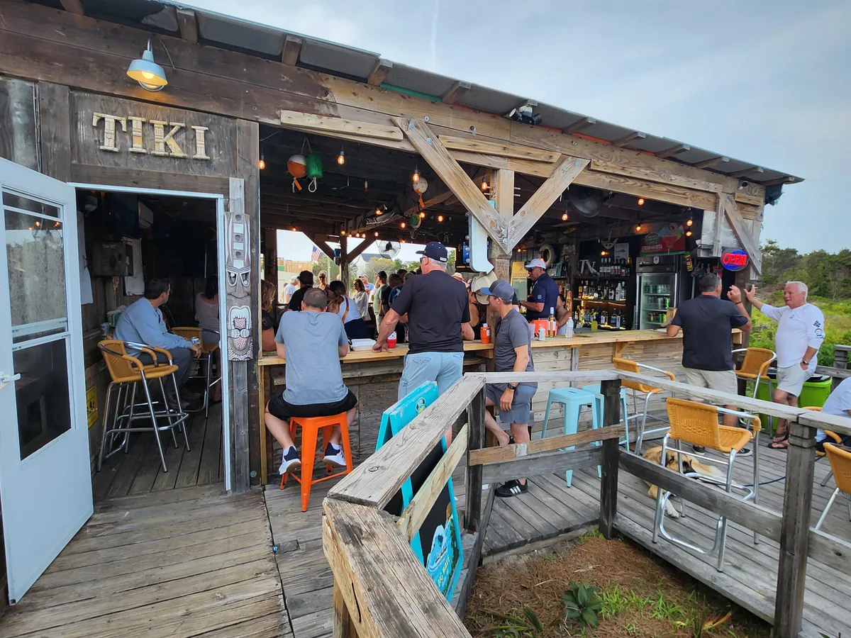 A lively exterior shot featuring a bright TIKI sign, a bustling bar area, and people socializing on the deck. The sunset glow and casual crowd suggest an inviting spot for drinks and casual dining, emphasizing the vibrant local dining scene.