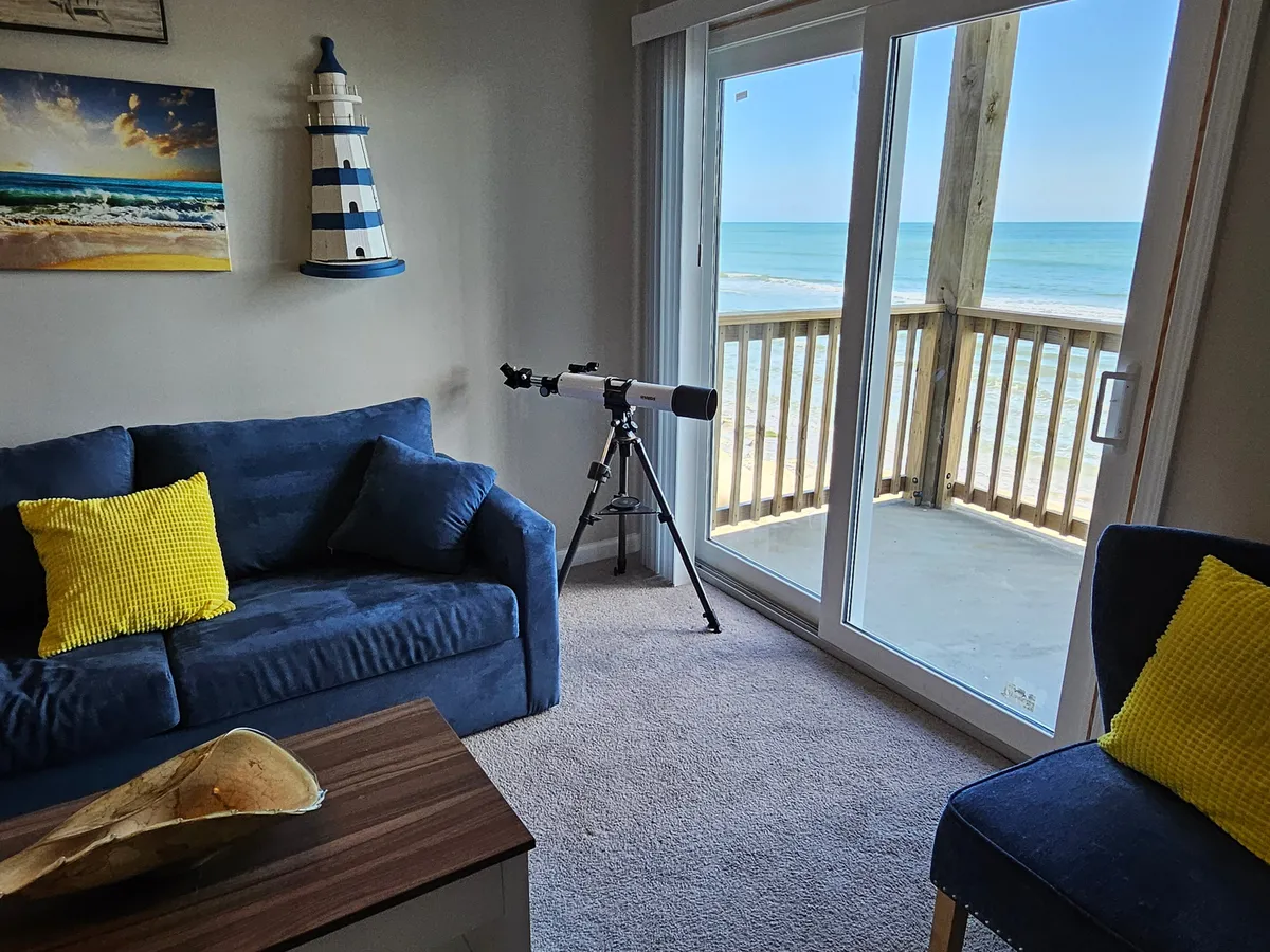 A tranquil and light-filled living area featuring a cozy sofa and a telescope by the window, with coastal lighthouse-themed decor and artwork. Large windows frame a spectacular view of the ocean, seamlessly blending indoor comfort with the beauty of the beach outside.
